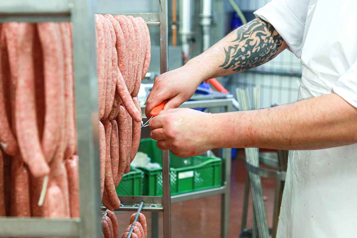 Zuschnitt von frischen Würsten in einer professionellen Wurstküche, Arbeiter mit Messer teilt Wurststränge in Portionen, Edelstahl-Arbeitstisch, hygienische Lebensmittelproduktion, handwerkliche Wurstverarbeitung, präziser Wurstzuschnitt.