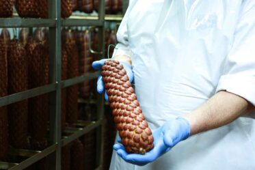 Wurstqualitätsprüfung in einer Lebensmittelproduktion, Fachkraft kontrolliert Farbe, Textur und Frische von Würsten auf einem Edelstahl-Arbeitstisch, hygienische Umgebung, Qualitätskontrolle in der Fleischverarbeitung, Prüfung von Wurstwaren nach Lebensmittelstandards.