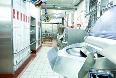 Innenaufnahme einer professionellen Wurstküche mit Edelstahl-Arbeitstischen, industriellen Fleischverarbeitungsmaschinen, hygienischen weißen Fliesen, Wurstfüllgerät, Fleischwolf und gekacheltem Boden; moderner lebensmittelverarbeitender Betrieb, hygienische Produktionsumgebung.