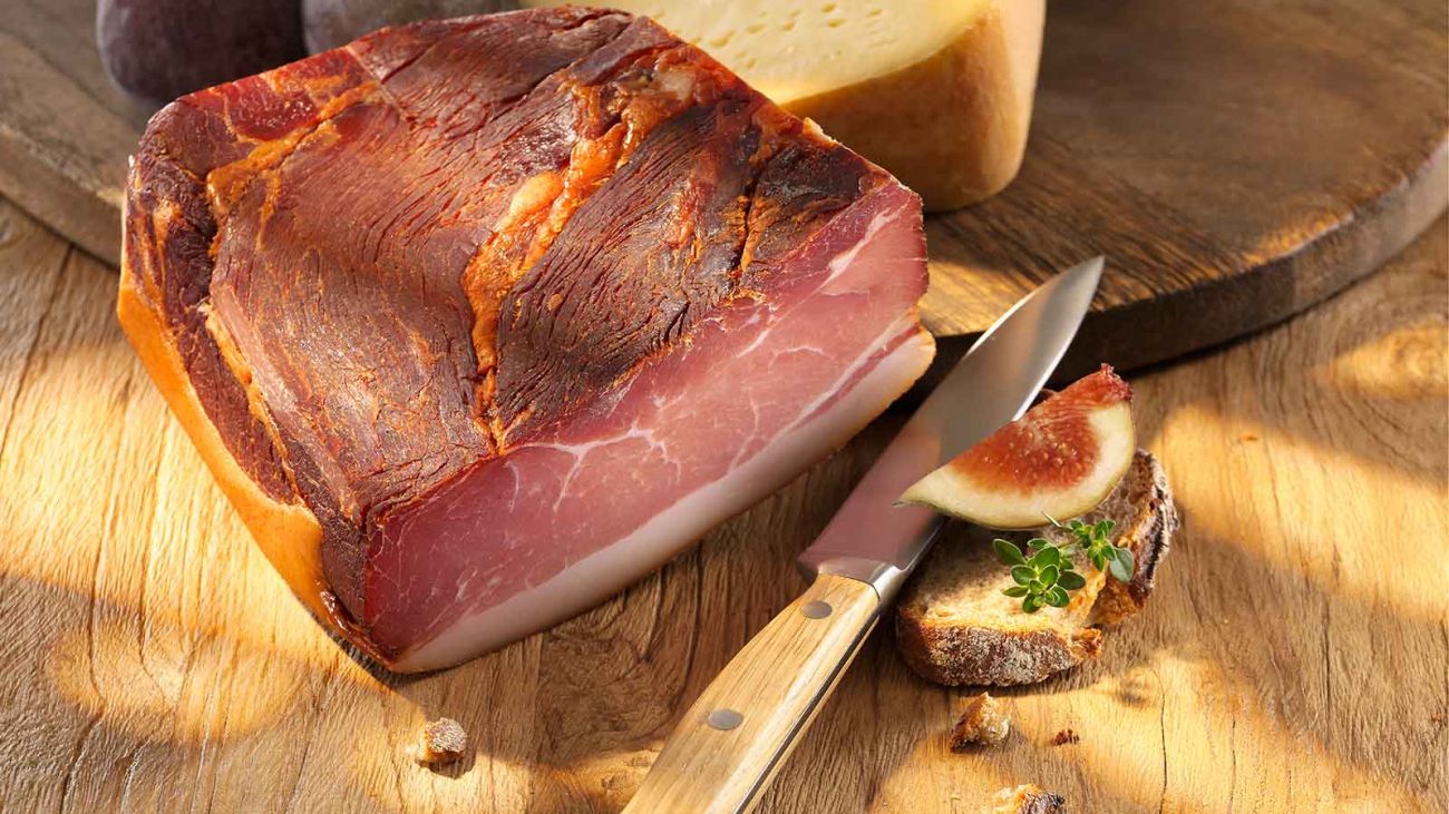 Ein Holzbrett mit Schinken und Messer, appetitlich präsentiert und umgeben von frischen Kräutern.  