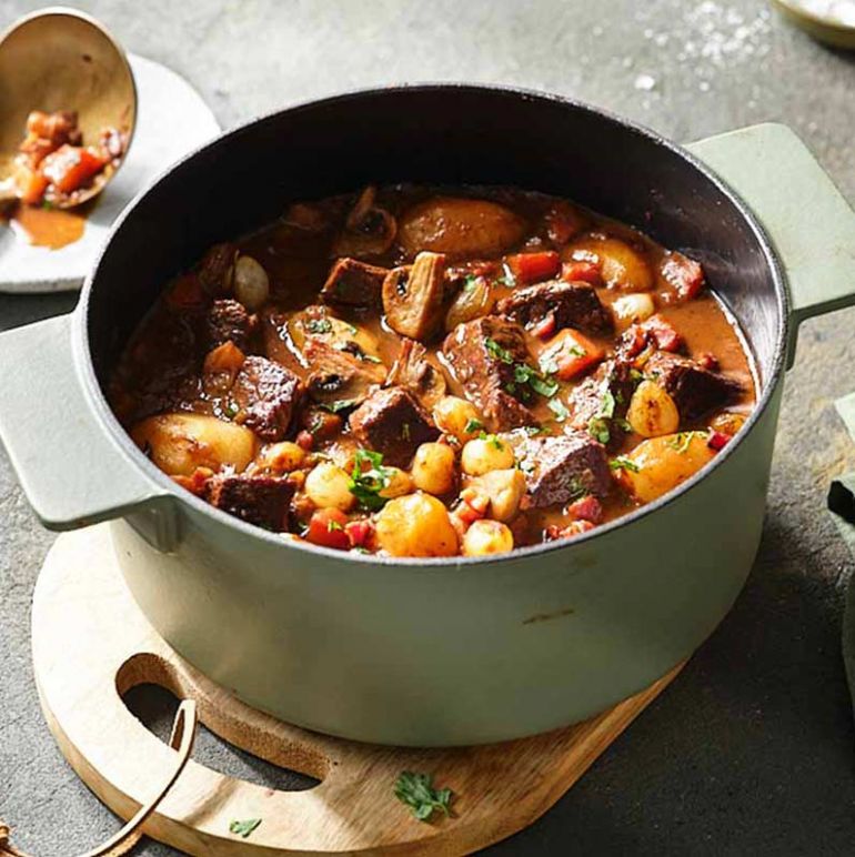 Ein rustikaler Topf mit Boeuf Bourguignon auf einem Holzbrett, dekoriert mit frischen Kräutern und Gewürzen.